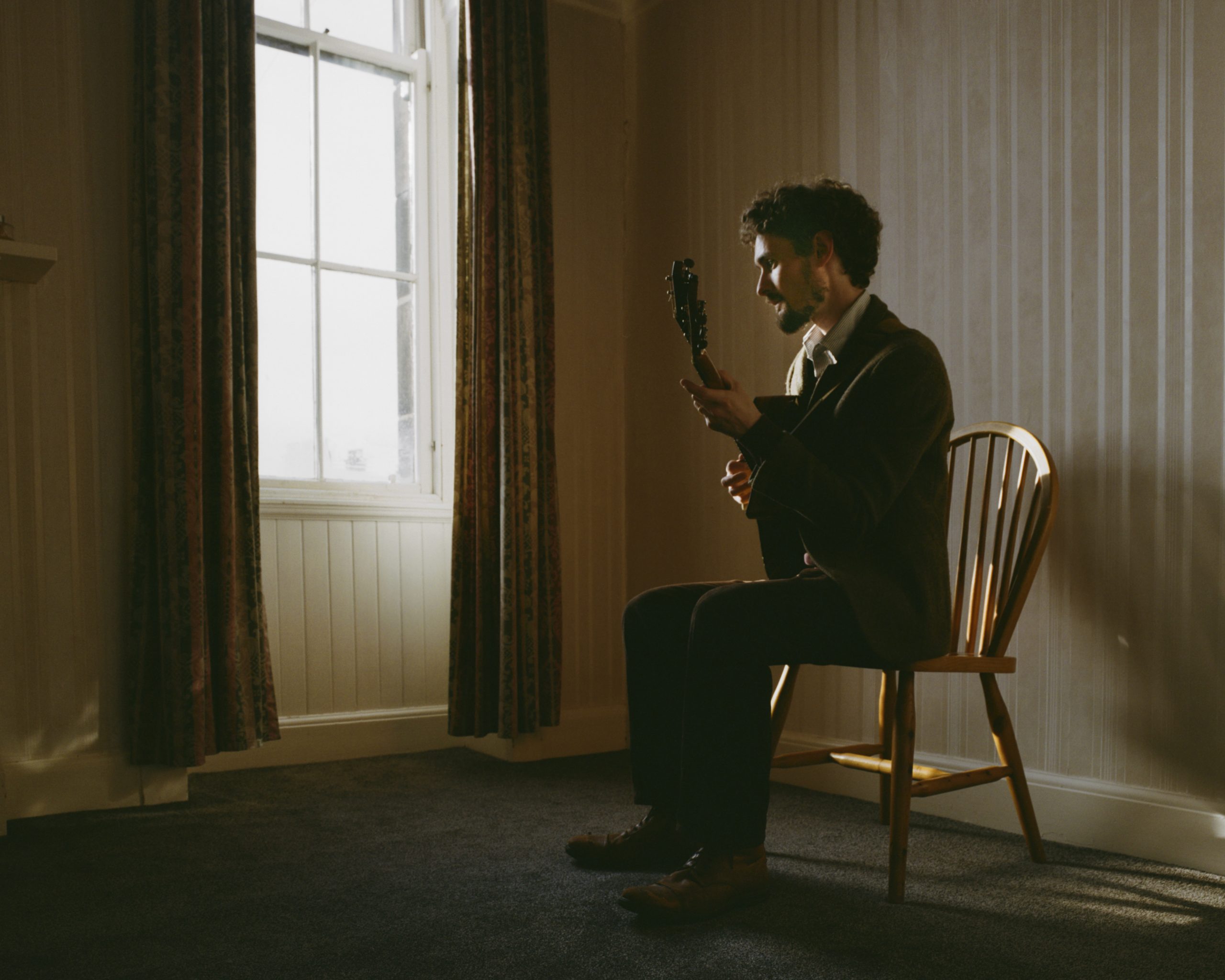 Chris Amer sits on a wooden chair with his guitar looking at a window, with soft lighting and curtains.