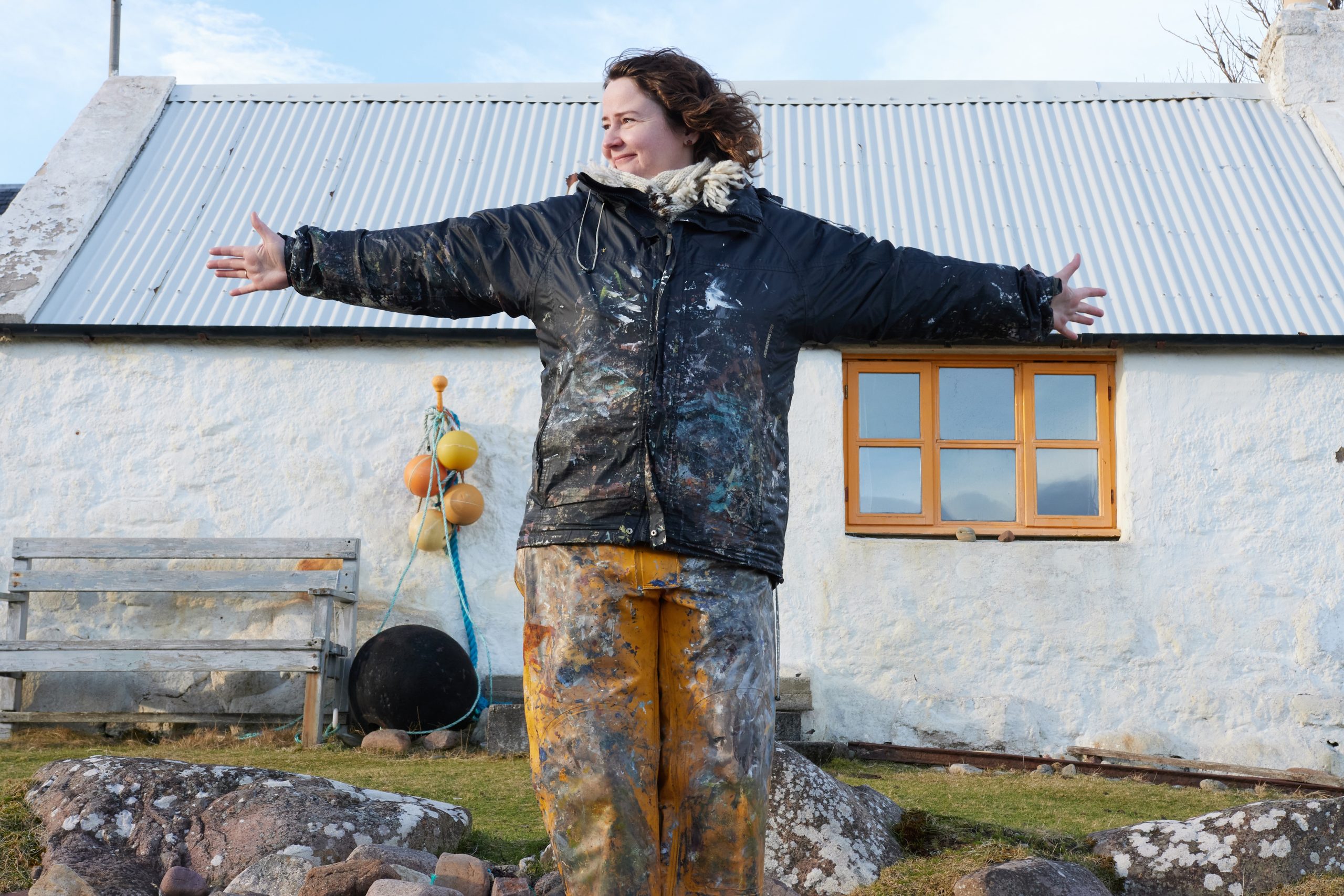 Mairearad Green stands smiling with her arms stretched out wearing yellow trousers and a black jacket covered in paint, in front of a little white cottage.